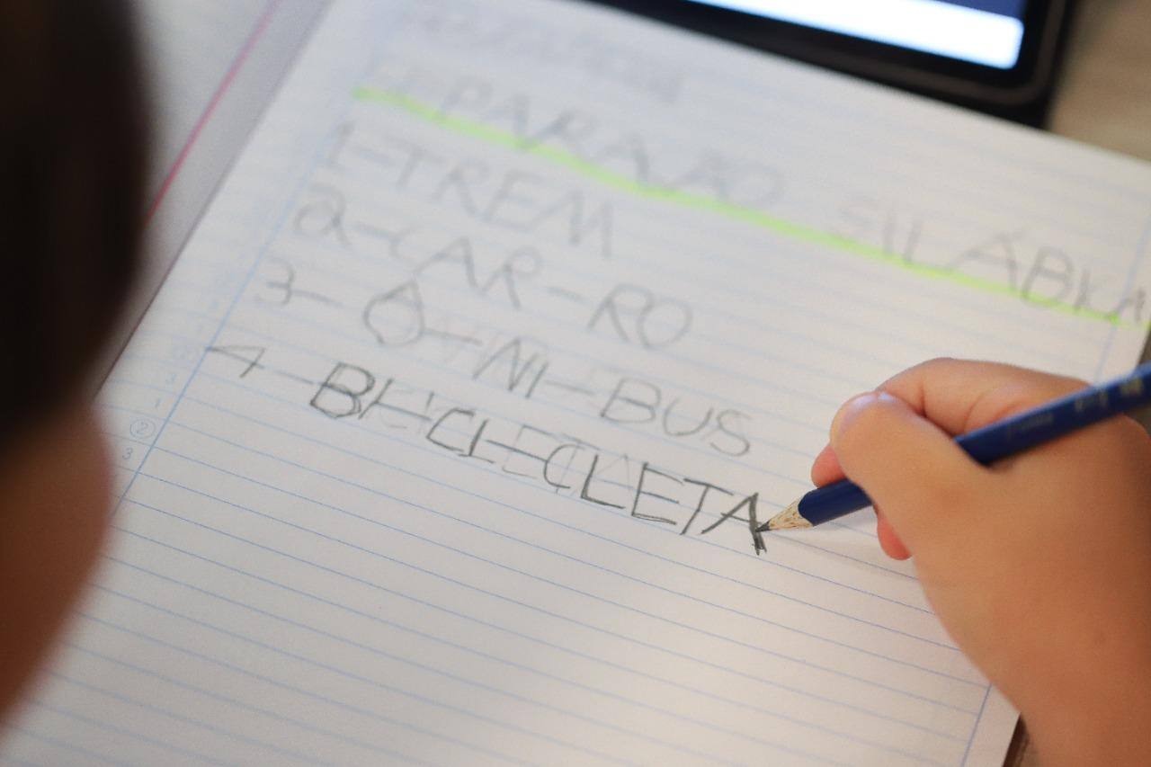 Imagem mostra criança aprendendo a escrever, com um lápis, em um caderno
