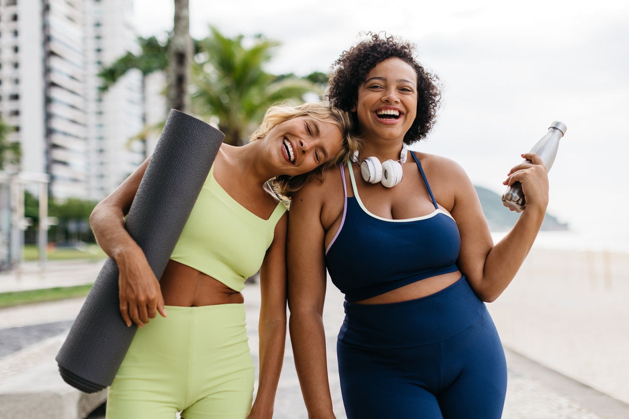 Portrait of two fitness friends smiling at the camera near the ocean, standing together with their yoga equipment in hand. Happy young women with different body sizes embracing an active lifestyle.