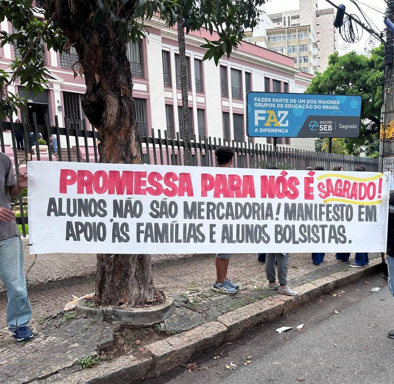 Foto mostra manifestação contra o fim das bolsas; pais seguram faixa escrita: "Promessa para nós é sagrado! Alunos não são mercadoria! Manifesto em apoio às famílias e alunos bolsistas"