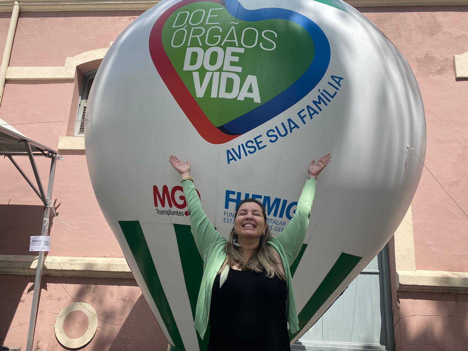 Foto mostra Ailla Pacheco, de 33 anos, que realizou um transplante de fígado em 2020, em frente a um balão da campanha lançada hoje