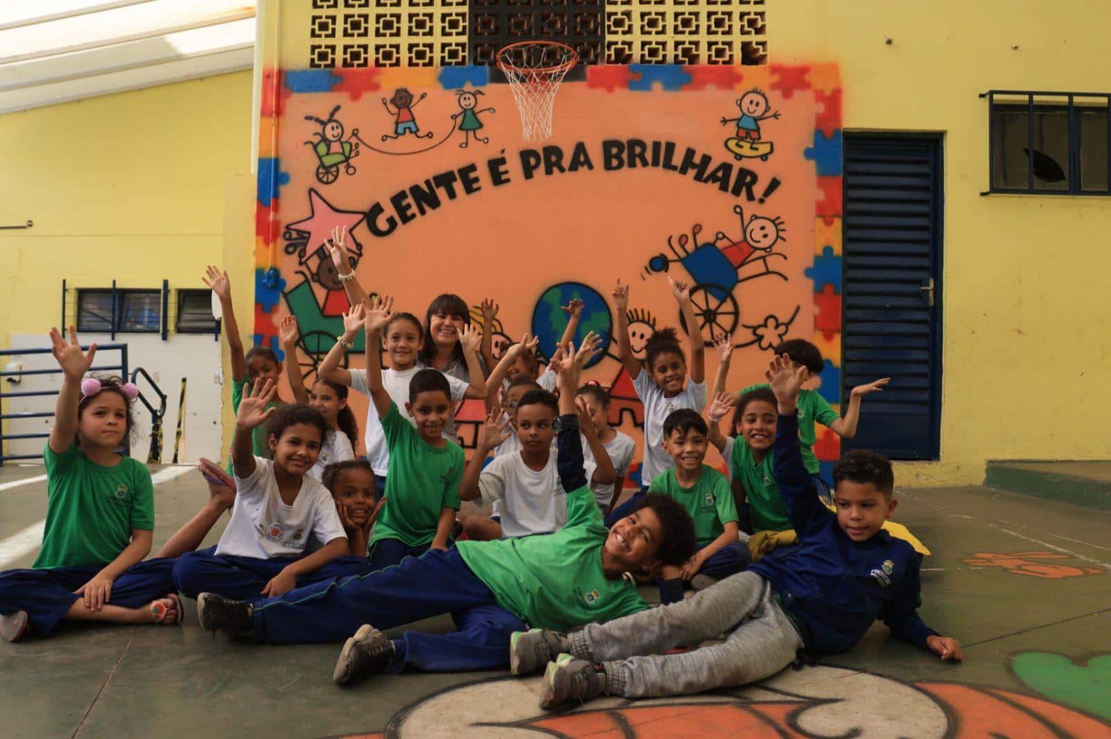Foto mostra professora Flor e seus alunos de 6 anos em frente a um mural que diz "Gente é pra brilhar"