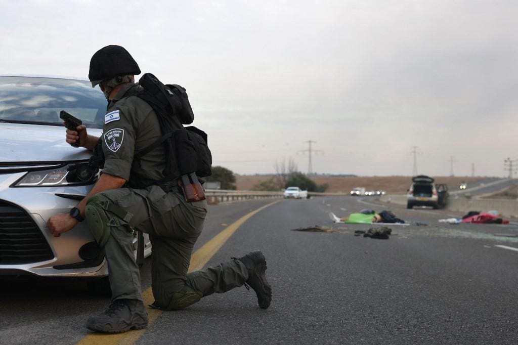 Um soldado israelense se esconde atrás de um carro enquanto olha para corpos em uma estrada principal perto do Kibutz Gevim, perto da fronteira com Gaza, em 7 de outubro de 2023
