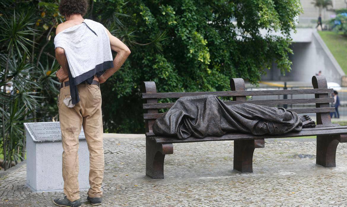 Pessoa em situação de rua lendo uma placa de uma escultura