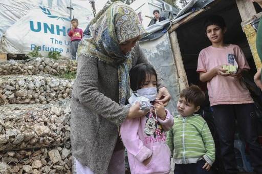 Uma mulher ajuda uma criança com uma máscara depois que membros da ONG "Team Humanity" distribuíram máscaras protetoras artesanais para migrantes e refugiados no campo de Moria, na ilha de Lesbos Uma mulher ajuda uma criança com uma máscara depois que membros da ONG "Team Humanity" distribuíram máscaras protetoras artesanais para migrantes e refugiados no campo de Moria, na ilha de Lesbos