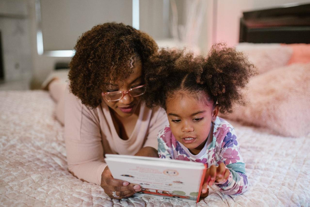 Imagem mostra mãe e filha, negras, deitadas em uma cama e lendo em um caderno