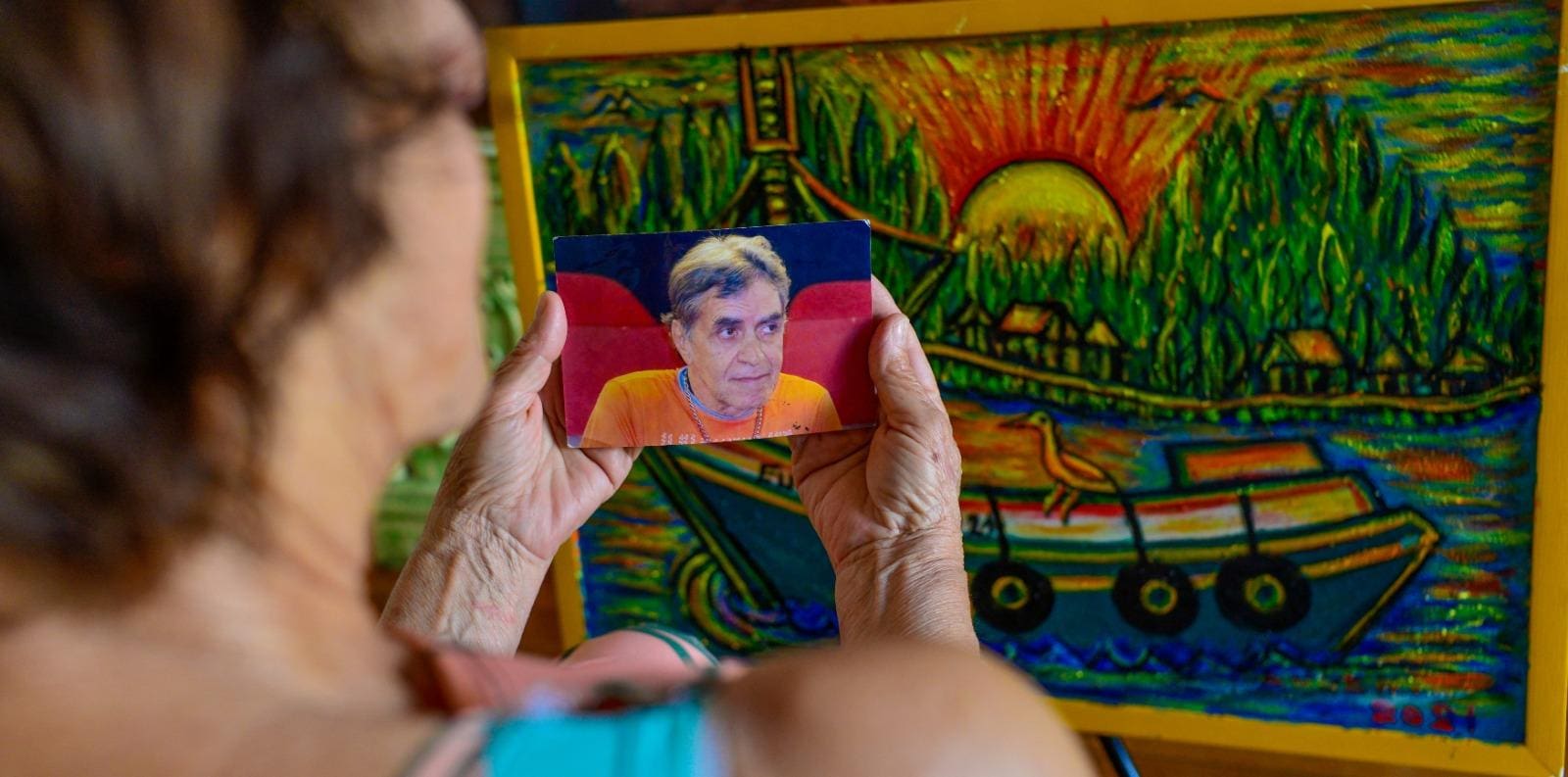 Maria Suzana, irmã de Queiroz, segura uma foto do homem que ficou 18 anos preso injustamente, em frente a um dos muitos quadros pintados por ele